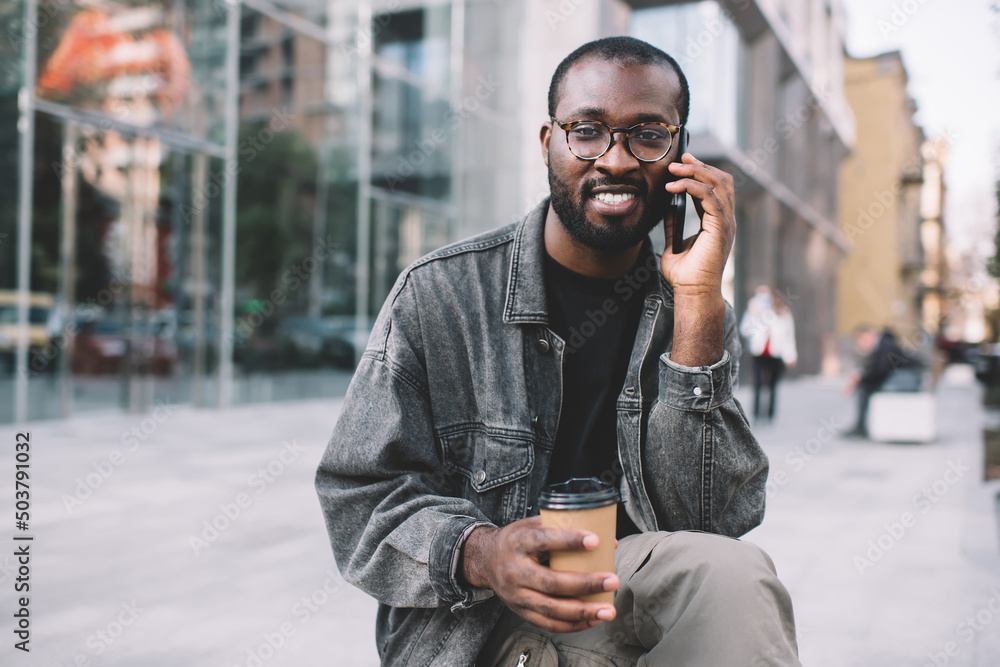 Portrait of dark skinned man in classic eyewear looking at camera while cell talking in urban city, millennial African American hipster guy with takeaway coffee using mobile technology for calling Portrait of dark skinned man in classic eyewear looking at camera while cell talking in urban city, millennial African American hipster guy with takeaway coffee using mobile technology for calling