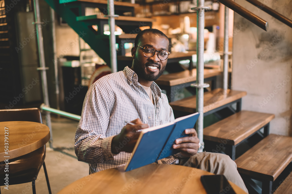 Portrait of happy student in eyewear holding copybook for creating education essay story, cheerful African American male with notebook for organization - learning and studying in coworking space Portrait of happy student in eyewear holding copybook for creating education essay story, cheerful African American male with notebook for organization - learning and studying in coworking space