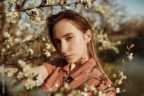 Portrait gorgeous young blonde woman in surrounded by flowering tree branches. Fashinable and stylish teenage girl looking at camera with calm and imperturbable poker face