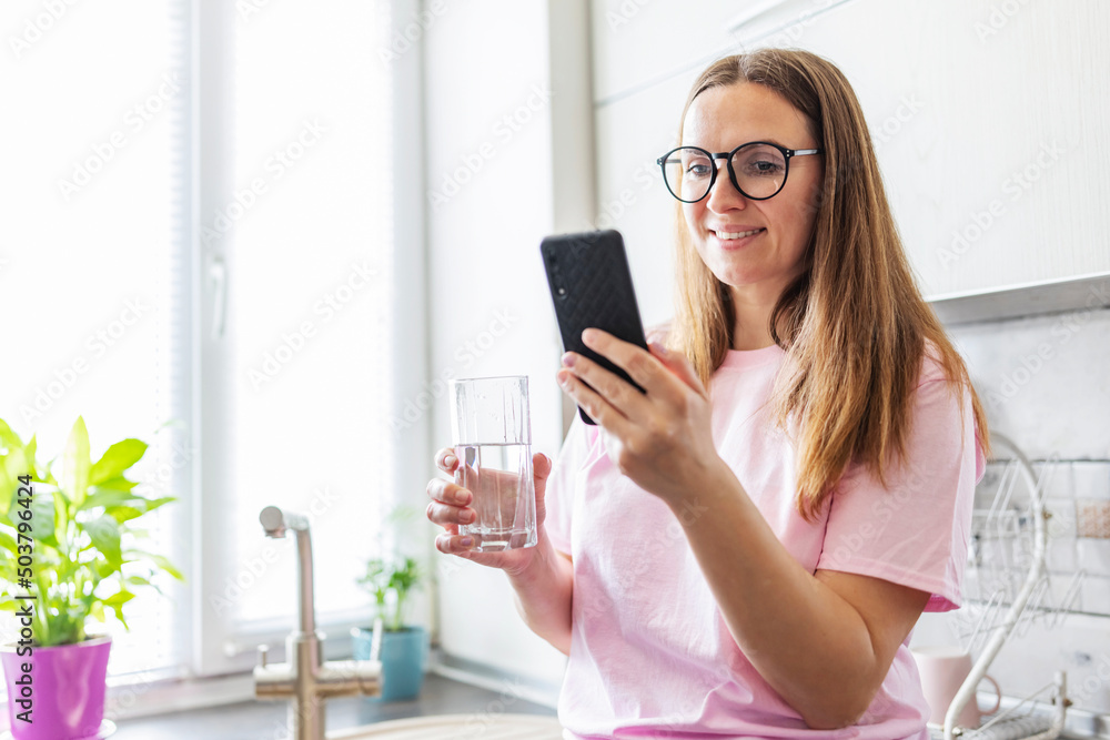 Beautiful young woman in eyeglasses drinking water while using mobile phone at home kitchen. Brunette girl reading or typing a text message Beautiful young woman in eyeglasses drinking water while using mobile phone at home kitchen. Brunette girl reading or typing a text message