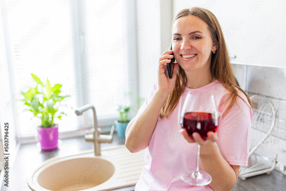 Happy relaxed young woman standing in kitchen with glass of red wine and using her smartphone