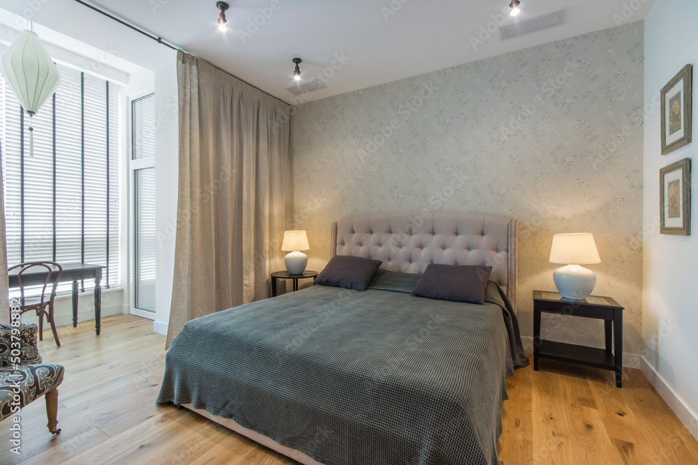 The interior of the bedroom with a panoramic window with blinds. A large bed with a gray blanket and an armchair. The interior of the bedroom with a panoramic window with blinds. A large bed with a gray blanket and an armchair.