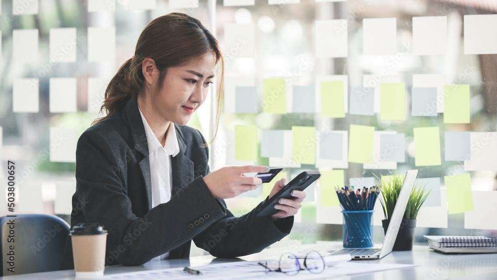 Asian woman holding a credit card and using a smartphone Businesswoman working at modern office, online shopping, ecommerce, internet banking, spending money, credit card working concept. Asian woman holding a credit card and using a smartphone Businesswoman working at modern office, online shopping, ecommerce, internet banking, spending money, credit card working concept.