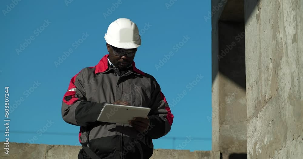 African american builder in white hard hat stands at construction site and makes notes in his Tablet PC. Cinema 4K 60fps video African american builder in white hard hat stands at construction site and makes notes in his Tablet PC. Cinema 4K 60fps video