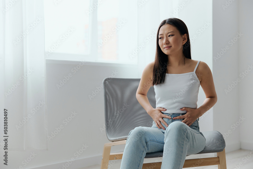 Stomach ulcer Gastritis. Tormented suffering tanned beautiful young Asian woman hold hands on stomach at home interior living room. Injuries Poor health Illness concept. Cool offer Banner Stomach ulcer Gastritis. Tormented suffering tanned beautiful young Asian woman hold hands on stomach at home interior living room. Injuries Poor health Illness concept. Cool offer Banner