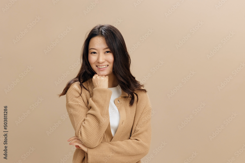 Cheerful happy smiling cute Asian student young woman in warm sweater reclines on fist posing isolated on over beige pastel studio background. Cool fashion offer. Seasonal Sale Fashion concept Cheerful happy smiling cute Asian student young woman in warm sweater reclines on fist posing isolated on over beige pastel studio background. Cool fashion offer. Seasonal Sale Fashion concept