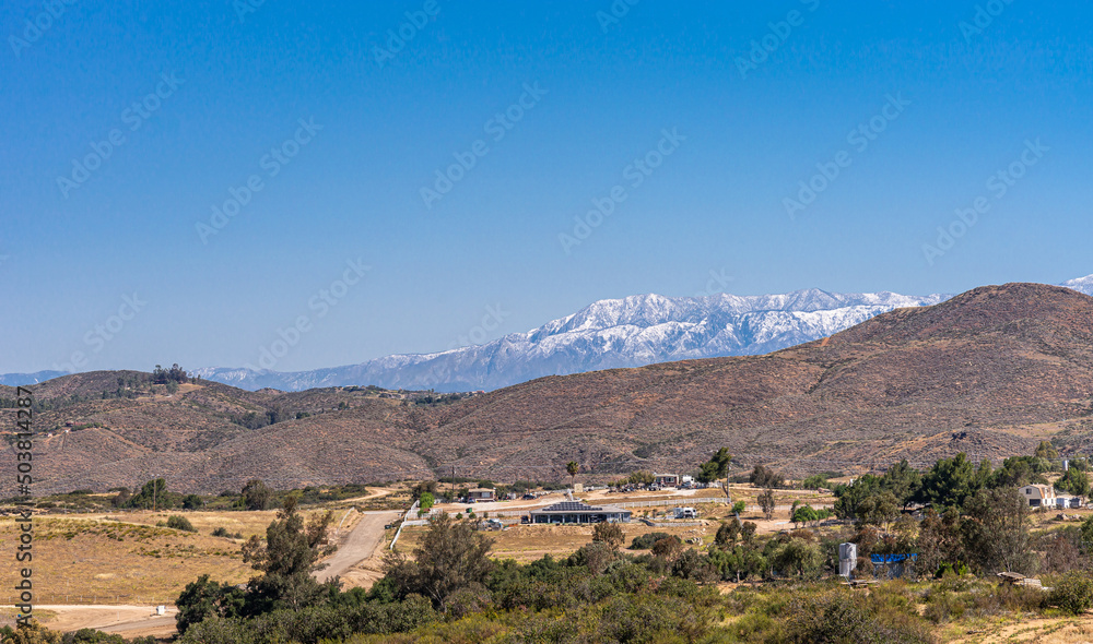 Temecula, CA, USA - April 23, 2022: Snow covered western part of San Jacinto mountains viewed from rural area north of Temecula under blue sky, with brown hills and ranches up front.