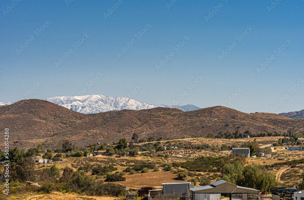 Temecula, CA, USA - April 23, 2022: Snow covered eastern part of San Jacinto mountains viewed from rural area north of Temecula under blue sky, with brown hills and ranches up front.