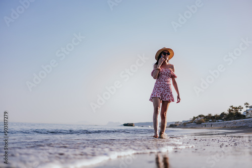 Brunette woman in red dress, hat and sunglasses talking happily with someone over phone, walking on beach