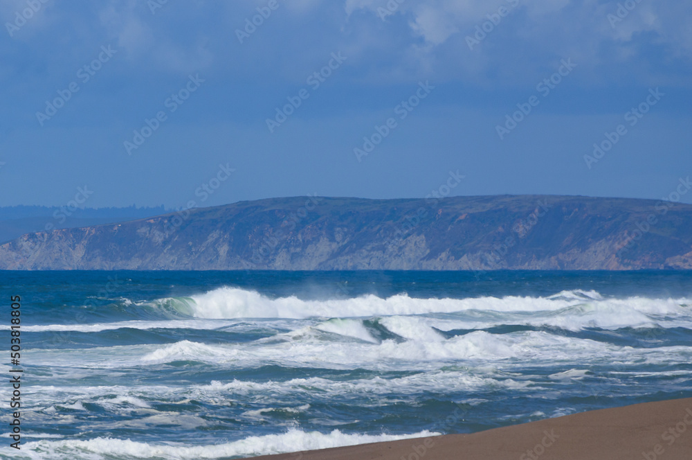 Fototapeta premium Crashing waves with mist rolling in to shore beautiful image for background with hills and bright blue sky