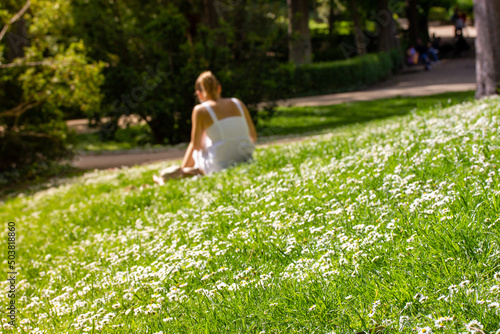A single young woman in a white dress, sundress lounging on a picnic on a green lawn on a sunny summer day. A girl in defocus resting in city park, on a blossoming meadow in day off. People and nature © vita