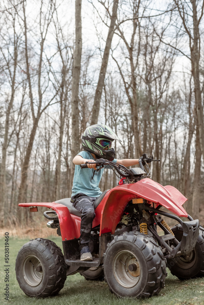 Boy riding a red four wheeler in grass with woods in background Boy riding a red four wheeler in grass with woods in background
