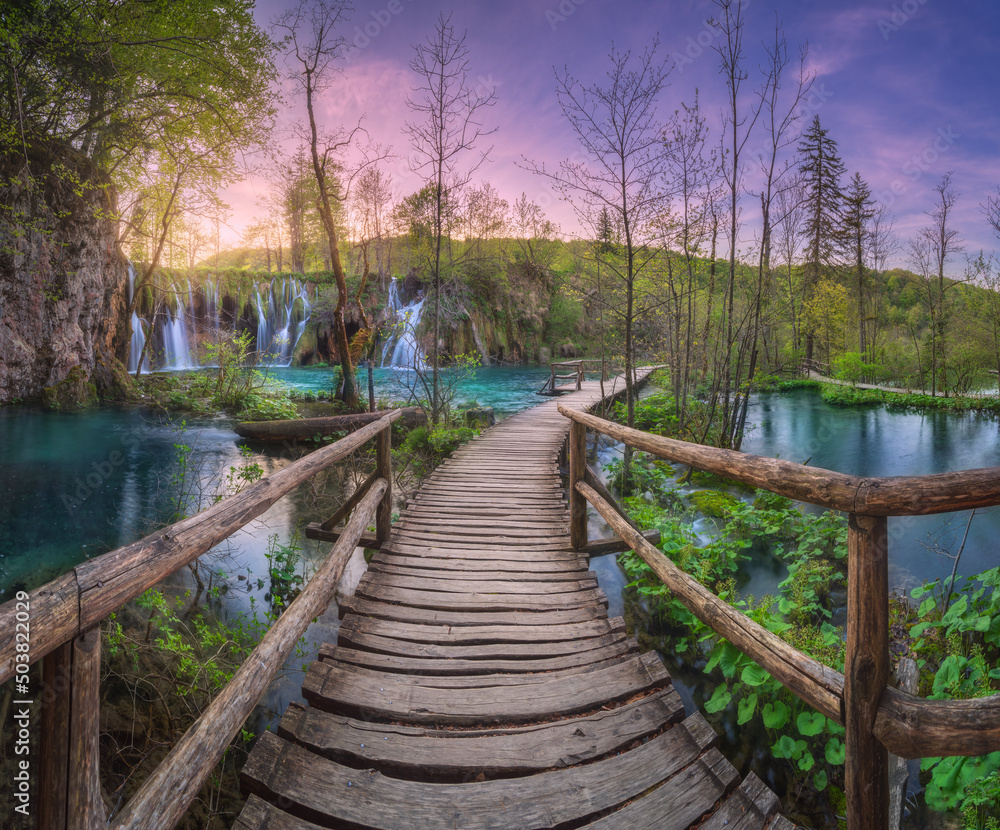 Waterfall and wooden path in green forest in Plitvice Lakes, Croatia at sunset in spring. Colorful landscape with trail in blooming park, trees, water lilies, river, pink sky in summer. Trail in woods Waterfall and wooden path in green forest in Plitvice Lakes, Croatia at sunset in spring. Colorful landscape with trail in blooming park, trees, water lilies, river, pink sky in summer. Trail in woods