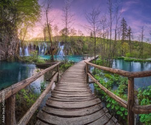 Waterfall and wooden path in green forest in Plitvice Lakes, Croatia at sunset in spring. Colorful landscape with trail in blooming park, trees, water lilies, river, pink sky in summer. Trail in woods