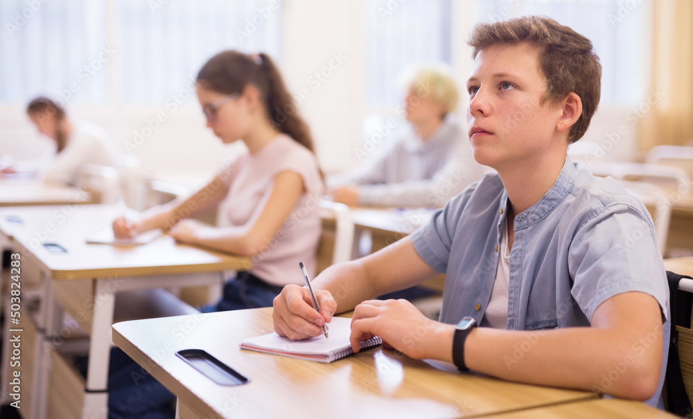 Fototapeta premium Teen boy sitting at desk in classroom full of pupils during lesson