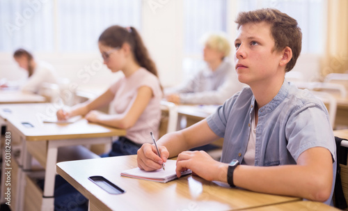 Teen boy sitting at desk in classroom full of pupils during lesson