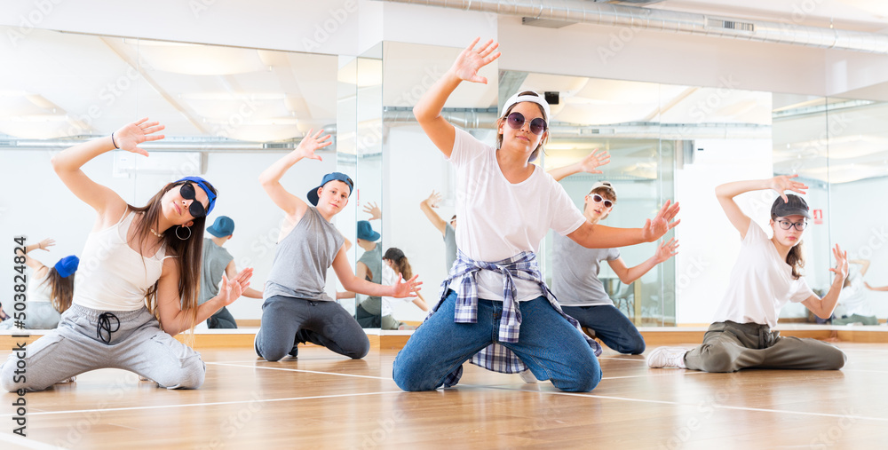 Positive teen girls and boys posing on knees in dance studio during hip hop class