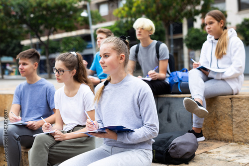 Attentive students write lesson in a notebook sitting on a stone street parapet