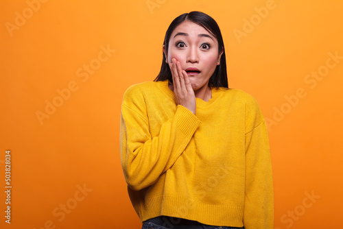 Shocked troubled asian woman wearing yellow vibrant sweater while putting hand on face. Uneasy young adult girl with shock expression, wearing vivid trendy clothing while on orange background. © DC Studio