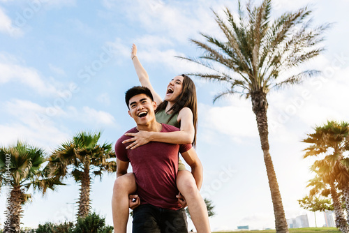 Happy young multiracial couple having fun together on summer holidays - Asian man piggybacking pretty woman outdoors - Love, happiness and friendship concept - Focus on woman face photo