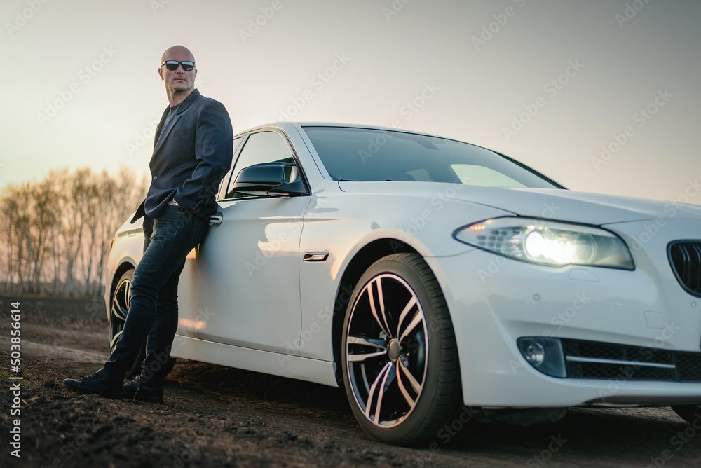 Young man in the suit stands near car on the dusty countryside road. Young man in the suit stands near car on the dusty countryside road.