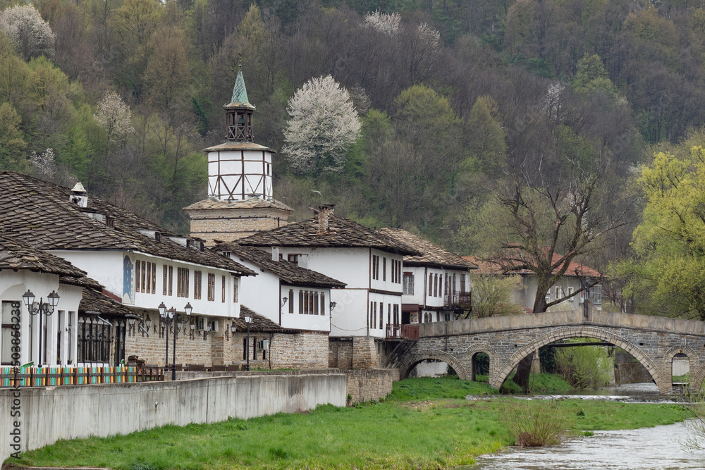 Old stone bridge and typical traditional Bulgarian houses with white walls, wooden windows, over the river and greenery. Historical architecture. Beautiful romantic rural exterior view of Tryavna town