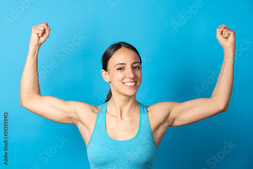 Smiling hispanic sportswoman showing biceps and looking at camera isolated on blue background.