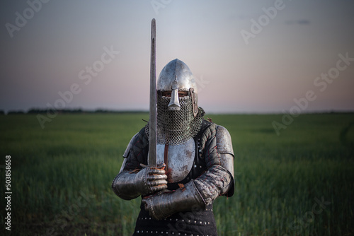 Knight guardian armor with a sword in hands stands in the field on a sunset background Fototapeta