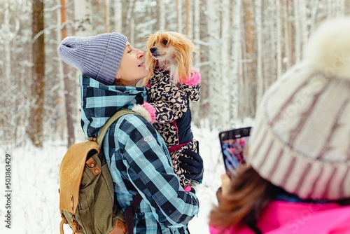 A teenager takes a photo on a smartphone of mom and dog. A woman is holding a Chinese Crested dog in her arms. Family walks outdoors in winter