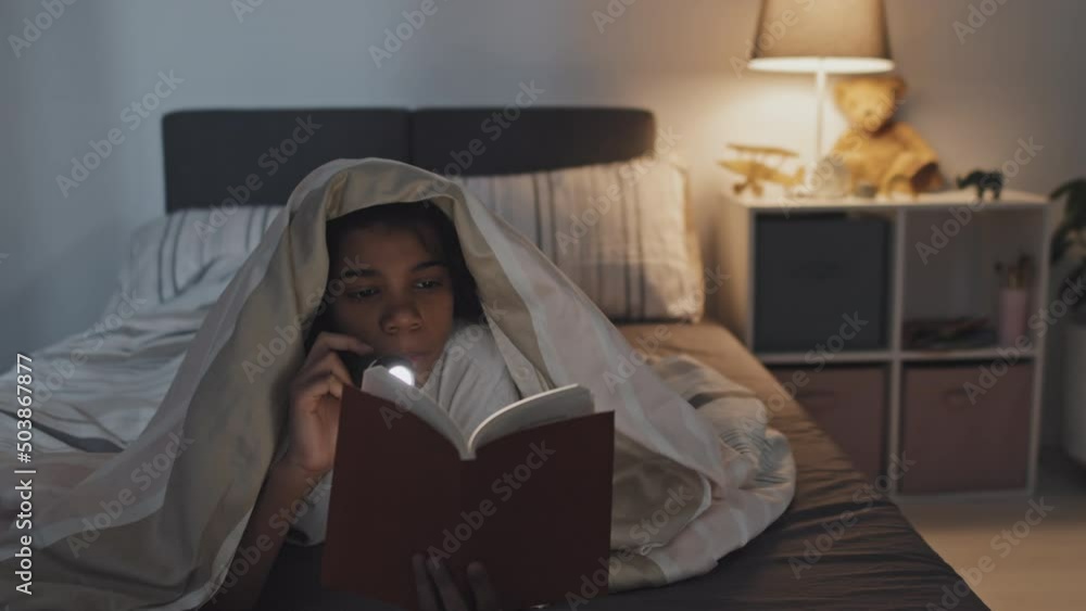 Medium close-up of twelve-year-old Black girl lying on stomach under blanket in bed, reading book using flashlight at night, yawning Medium close-up of twelve-year-old Black girl lying on stomach under blanket in bed, reading book using flashlight at night, yawning