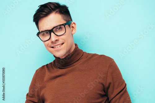 Portrait of handsome smiling model. Sexy stylish man dressed in brown turtleneck sweater and jeans. Fashion hipster male posing near blue wall in studio. In spectacles. Eyewear