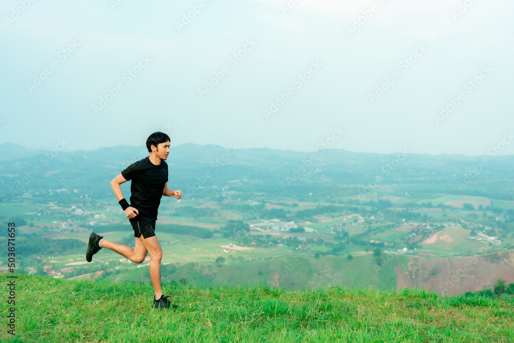 Young Asian runner, wearing black sportswear, running on a big mountain trail, cool morning, windmills, and sky in the background.. Young Asian runner, wearing black sportswear, running on a big mountain trail, cool morning, windmills, and sky in the background..
