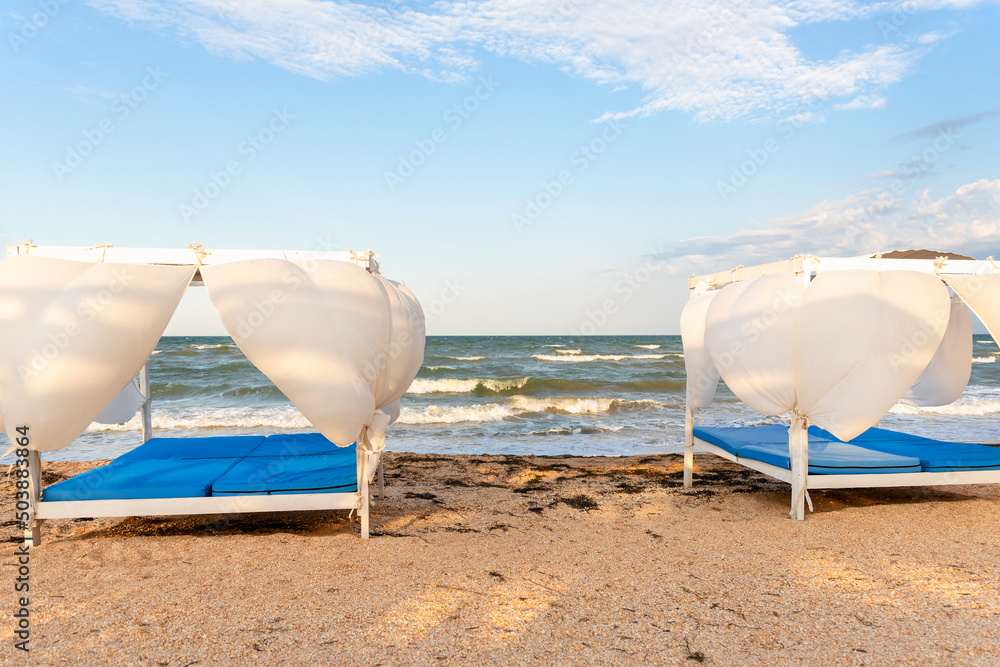 Scenic view empty white beach wooden canopies cabanas with white curtain blowing by wind against stormy sea or ocean san seaside. Luxury sunbed for spa and leisure vacation travel