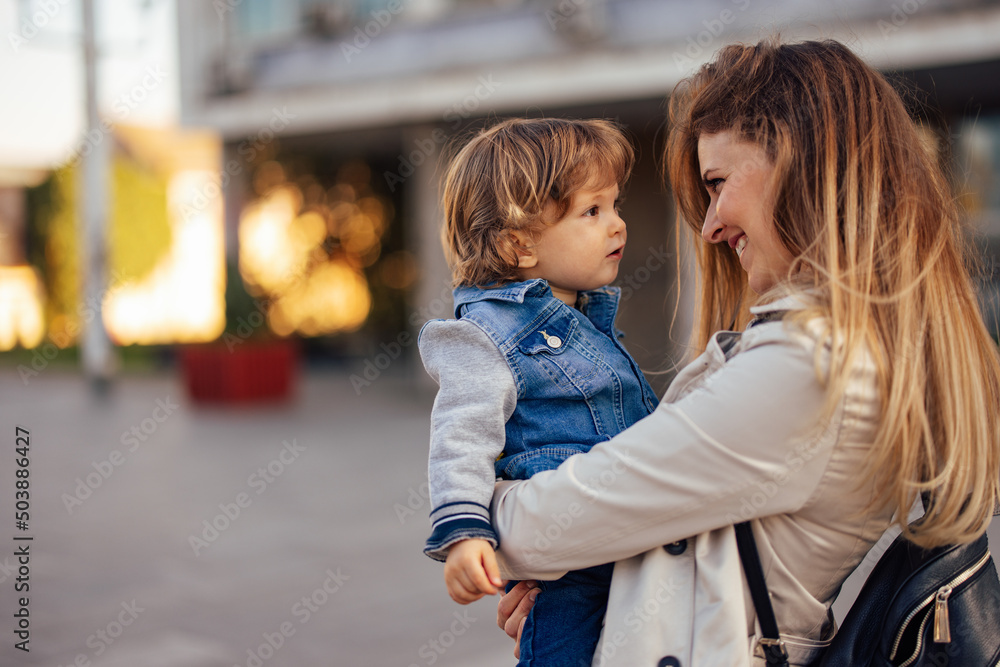 Blonde smiling mother, looking at her son while holding him with both hands outdoor. Blonde smiling mother, looking at her son while holding him with both hands outdoor.