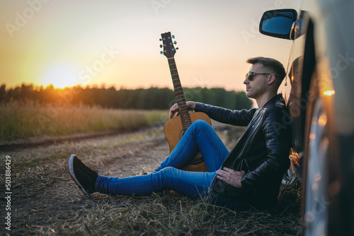 Musician with guitar sits on the dusty countryside road near the car in the sundown rays.