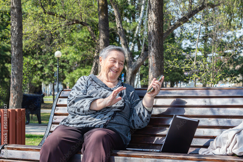Attractive senior woman sitting on bench using smartphone and computer for remote online chat.
