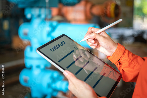 Action of a human is using digital pen to perform checklist on tablet with background of factory workplace. Industrial and technology working concept. Close-up and selective focus at hand's part.