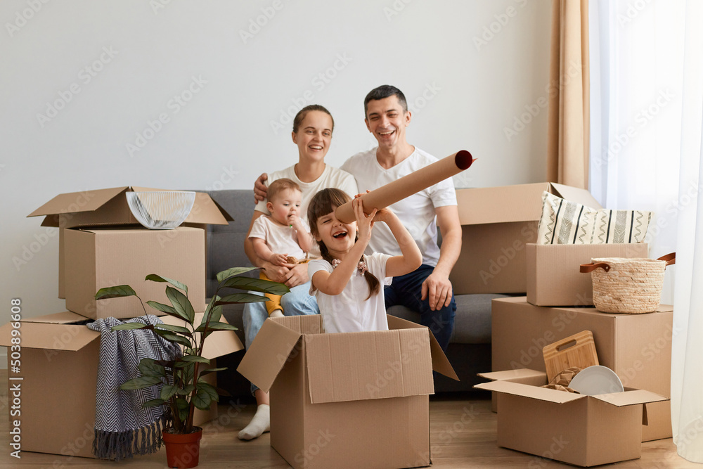 Horizontal shot of happy family relocating in new flat and having fun together while unpacking their personal belongings, little daughter sitting in cardboard box with handmade spyglass. Horizontal shot of happy family relocating in new flat and having fun together while unpacking their personal belongings, little daughter sitting in cardboard box with handmade spyglass.