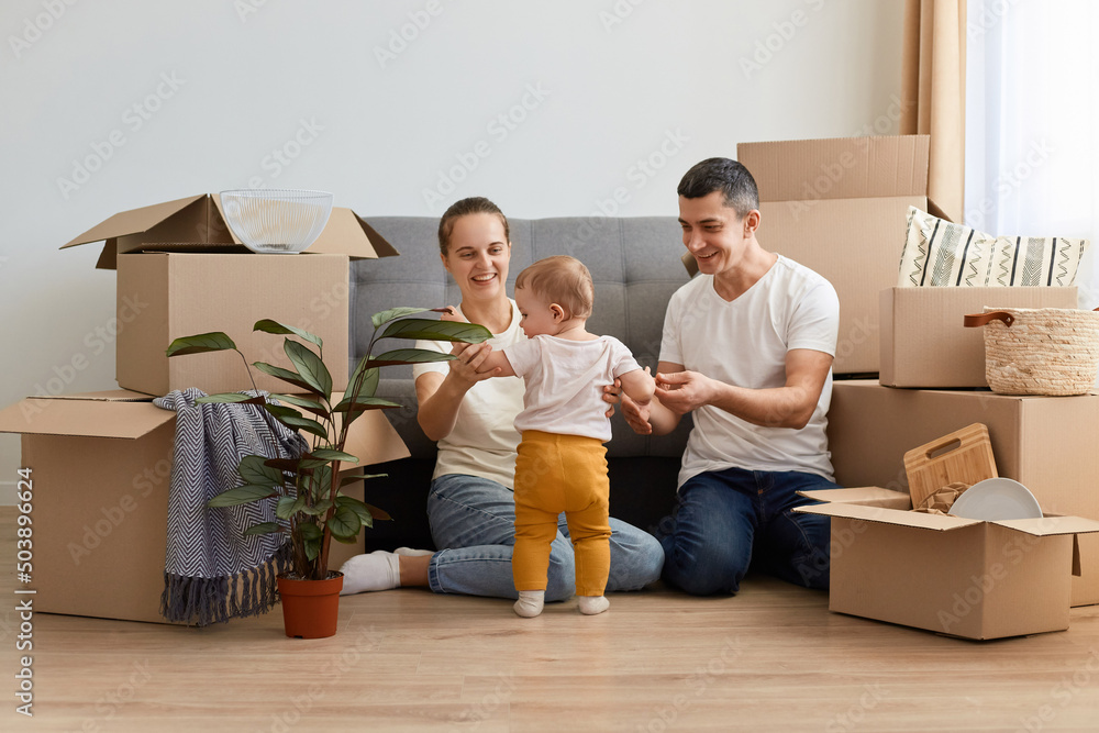 Portrait of young family unpacking moving box at their new apartment, sitting on floor near sofa and playing with their infant kid, smiling happily, relocating in a new home. Portrait of young family unpacking moving box at their new apartment, sitting on floor near sofa and playing with their infant kid, smiling happily, relocating in a new home.