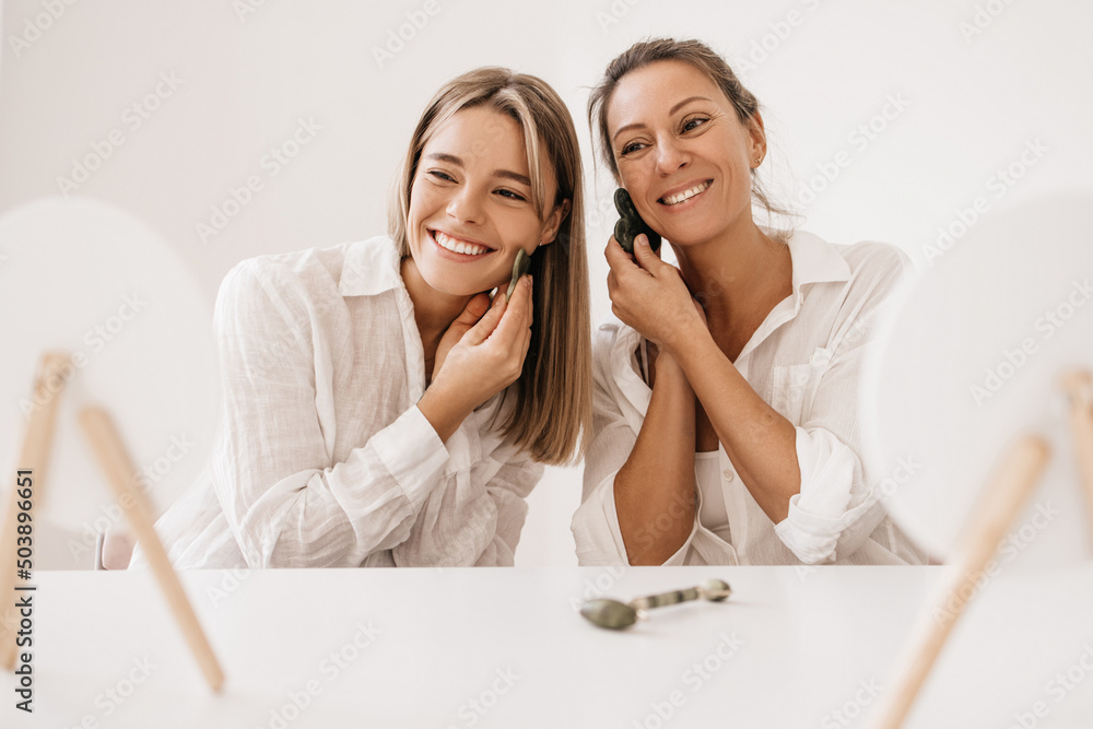 Young caucasian women doing facial massage with scraper in front of mirror on white background. Blondes wear shirts using gouache jade stone at home. Cosmetic products, concept Young caucasian women doing facial massage with scraper in front of mirror on white background. Blondes wear shirts using gouache jade stone at home. Cosmetic products, concept