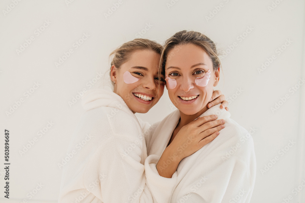 Positive two young caucasian women smile teeth standing inembrace on white background. Blondes wear bathrobes and patches during spa treatments. Concept of natural cosmetics, wrinkle smoothing - obrazy, fototapety, plakaty 
