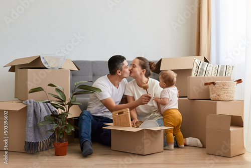 Portrait of couple sitting on floor near sofa among carton parcels with their baby girl and kissing, celebrating relocating in new house, expressing happiness and love.