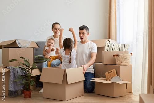 Indoor shot of beautiful positive optimistic family unpacking moving box at their new house and playing with cardboard boxes, couple with their children having fun while moving in new apartment.