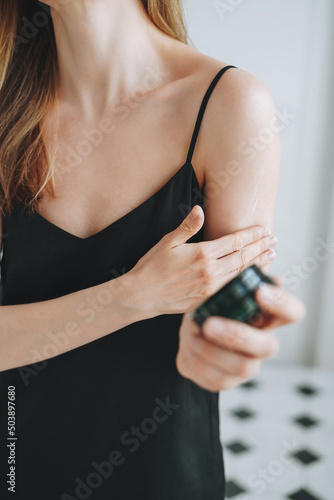 Young woman with dark long hair in black dress put day nourishing moisturizer body cream on clean fresh skin in bathroom at home