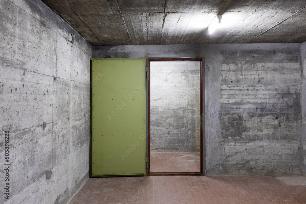 Front view of reinforced concrete wall of a bunker with open vault armored door. Scene illuminated by a white neon lamp