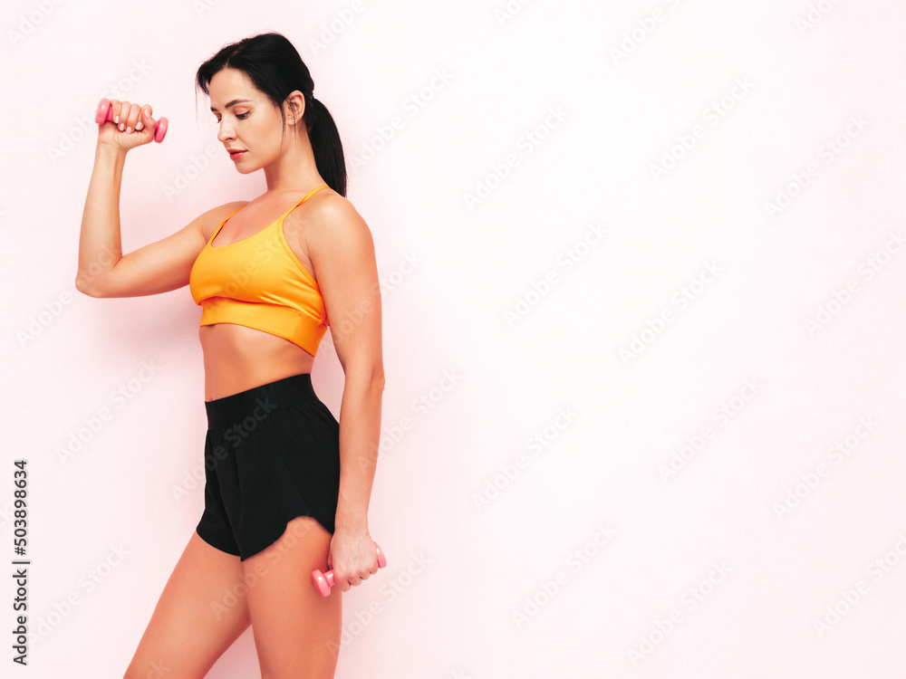 Fitness confident woman in black sports clothing. Sexy young beautiful model with perfect body. Female isolated on pink wall in studio. Stretching out before training.Making exercises with dumbbells Fitness confident woman in black sports clothing. Sexy young beautiful model with perfect body. Female isolated on pink wall in studio. Stretching out before training.Making exercises with dumbbells