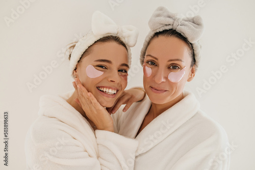 Close-up of caucasian young and adult women with patches under eyes look at camera on white background. Beauty wear bathrobes and hair bands. Skin care concept