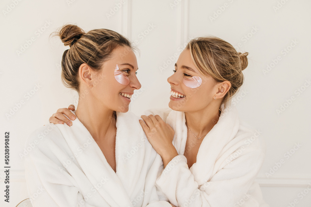 Attractive caucasian young girls in bathrobes are smiling looking at each other on white background. Blondes in good mood use patches. Beauty needs care concept - obrazy, fototapety, plakaty 