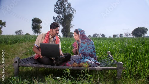 Young village couple in their green wheat fields in India - Using laptop sitting on a cot  modern villager. A beautiful housewife of Indian village knitting clothes while sitting on a Chaarpai - in... photo