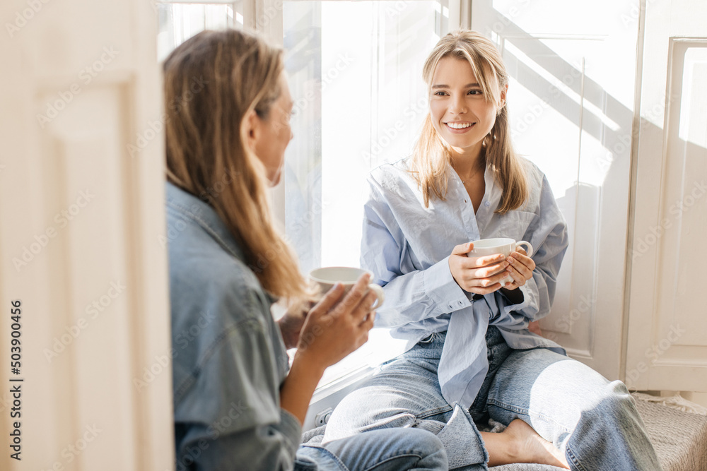 © Look! - Smiling young caucasian women drink delicious coffee and gossip sitting on windowsill in morning. Blondes wear casual blue clothes. Happy weekend concept © Look! - Smiling young caucasian women drink delicious coffee and gossip sitting on windowsill in morning. Blondes wear casual blue clothes. Happy weekend concept